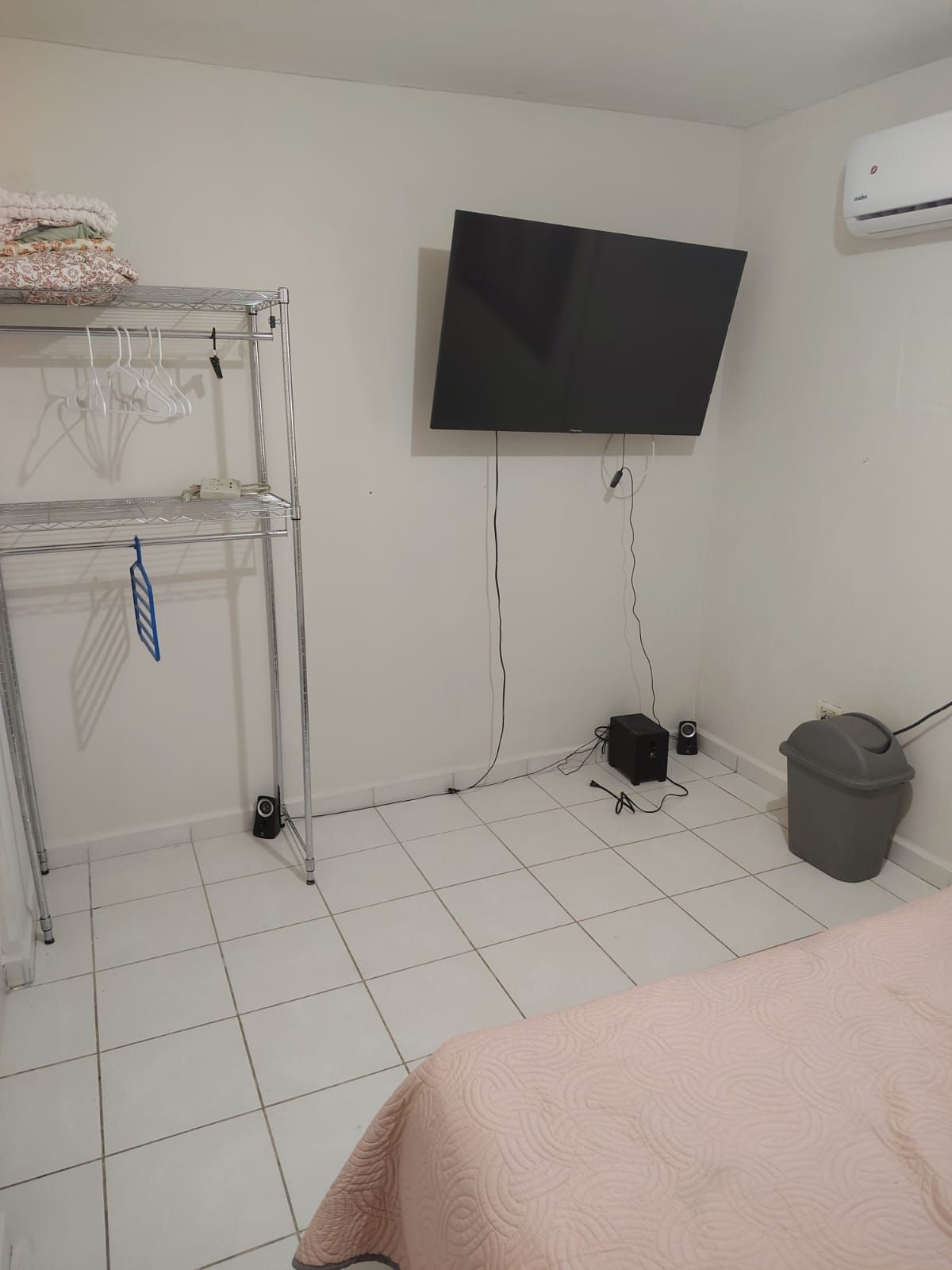 Une chambre avec un lit, un téléviseur fixé au mur, une étagère avec des cintres et une poubelle.