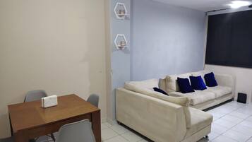Ein Zimmer mit einem Holztisch, zwei Stühlen, einem cremefarbenen Sofa mit blauen Kissen und einer weißen Wand mit zwei sechseckigen Regalen.