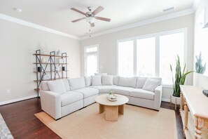 A modern living room with a sectional sofa, a round coffee table, a bookshelf, and a ceiling fan. - 4BR, 3.5BA beach retreat in Virginia Beach with parking, patio & kitchen. (Virginia Beach)