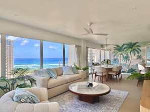 Ein modernes Wohnzimmer mit einer großen Couch, einem Couchtisch und einem Essbereich mit Stühlen. Große Fenster bieten Blick auf den Ozean und Palmen.