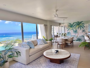 Ein modernes Wohnzimmer mit großem Fenster mit Blick auf einen Strand, einem runden Couchtisch und einem Essbereich mit Stühlen.