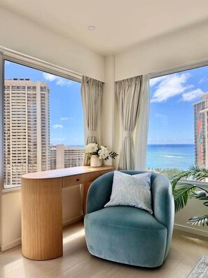 A modern room with a curved wooden desk, a blue armchair, and a view of tall buildings and the ocean. - 2-Bedroom 2-Bath Oceanfront House w/ Washer Dryer (Honolulu)