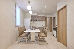 A modern dining area with a rectangular table, cushioned chairs, a hanging light fixture, and a kitchen in the background. - eni suite macchamachi (Chuo-ku)