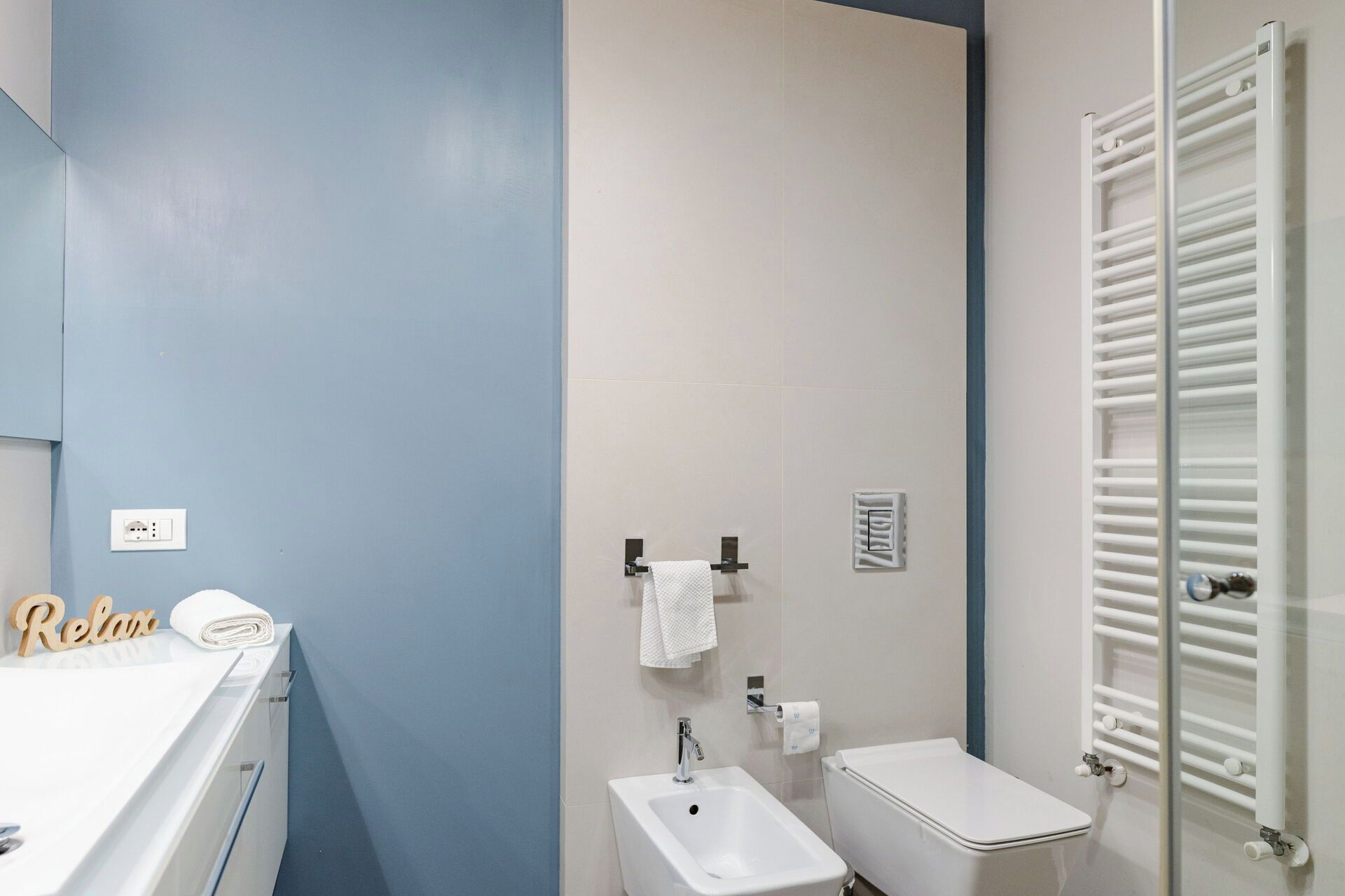 Een badkamer met een wastafel, bidet, toilet, handdoekrek en een handdoek met de tekst 'Relax'.