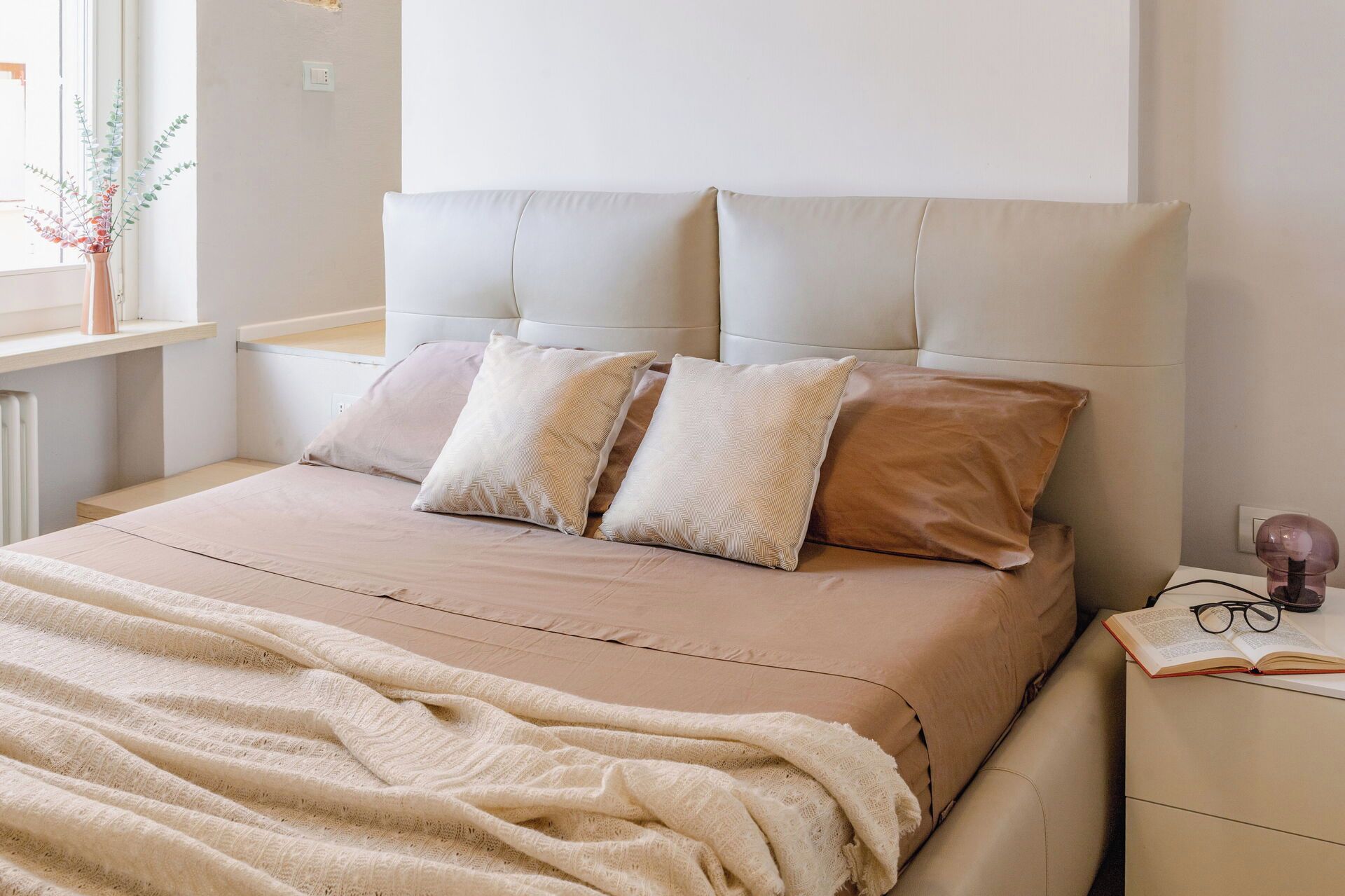 A bed with a headboard, multiple pillows, a blanket, and a bedside table with glasses and a book.