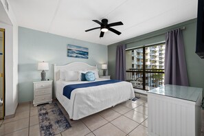 A hotel room with a bed, two bedside tables, a ceiling fan, a window with curtains, and a view of a building. - Saida III #606 | Beachfront Condo • 3 Pools • Tennis & Pickleball (South Padre Island)