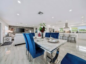 A modern kitchen with white cabinets, a central island with bar stools, and a dining area with blue chairs and a marble table. - Two-Story 3BR with Pool Table and Private Yard (St. Petersburg)