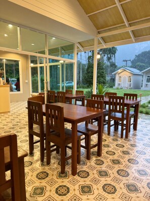 Un espacio de comedor al aire libre cubierto con suelo estampado, mesas y sillas de madera, y grandes ventanales que ofrecen vistas del paisaje circundante.