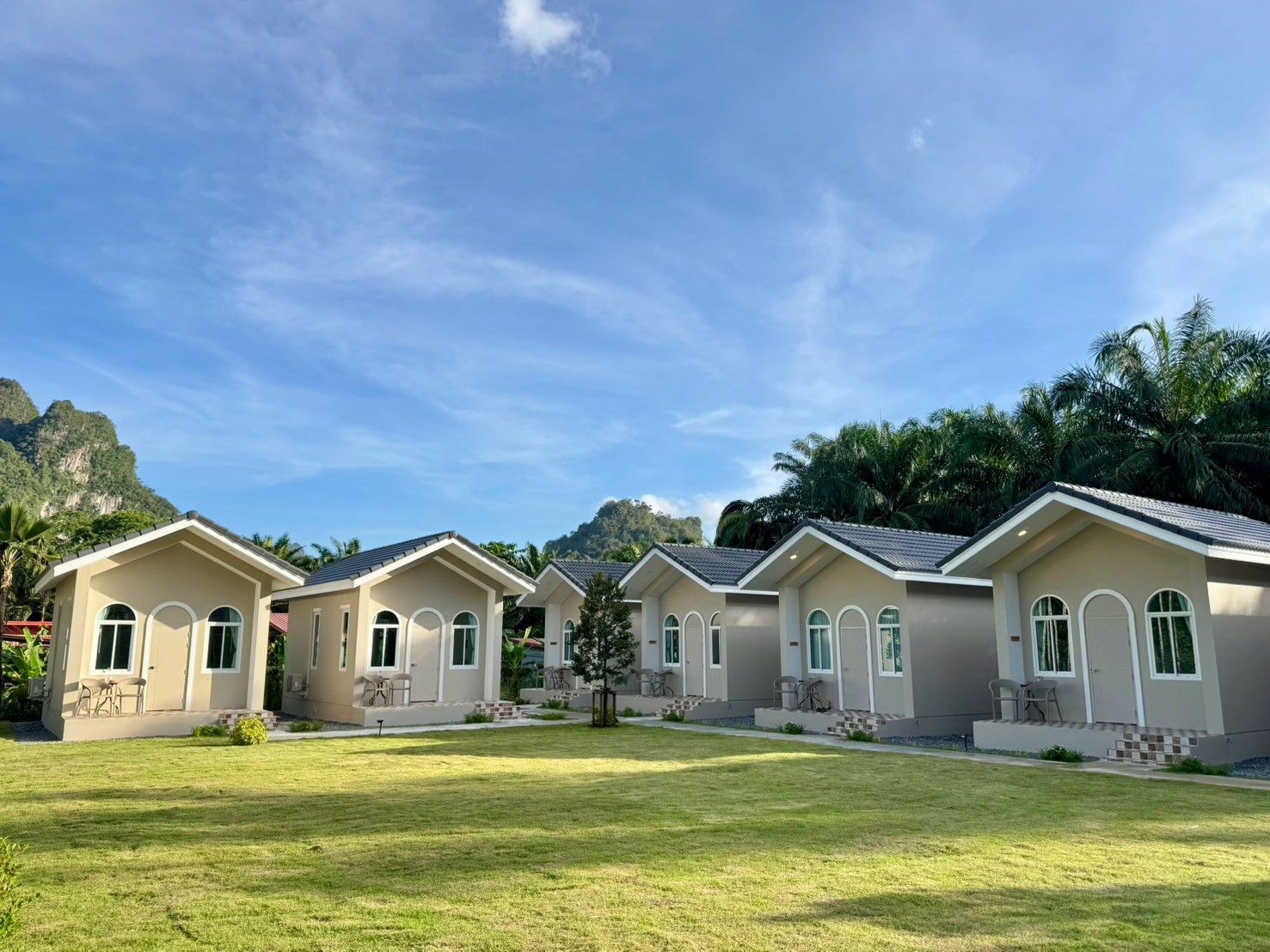 A row of modern, single-story buildings with large windows and a flat roof, surrounded by a well-maintained lawn and palm trees, with a mountainous backdrop.