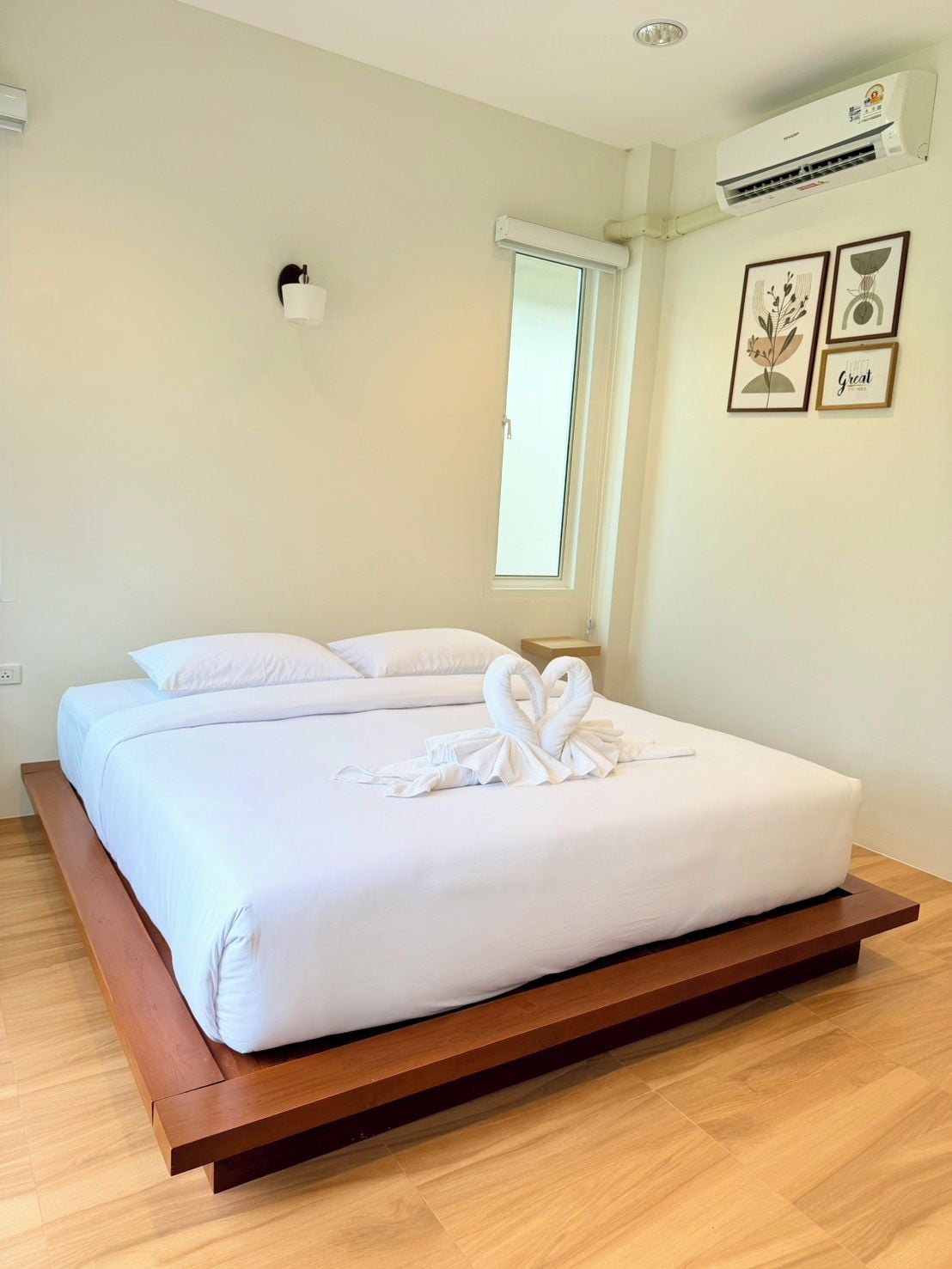 A hotel room with a bed, a window, an air conditioning unit, and framed artwork on the wall.