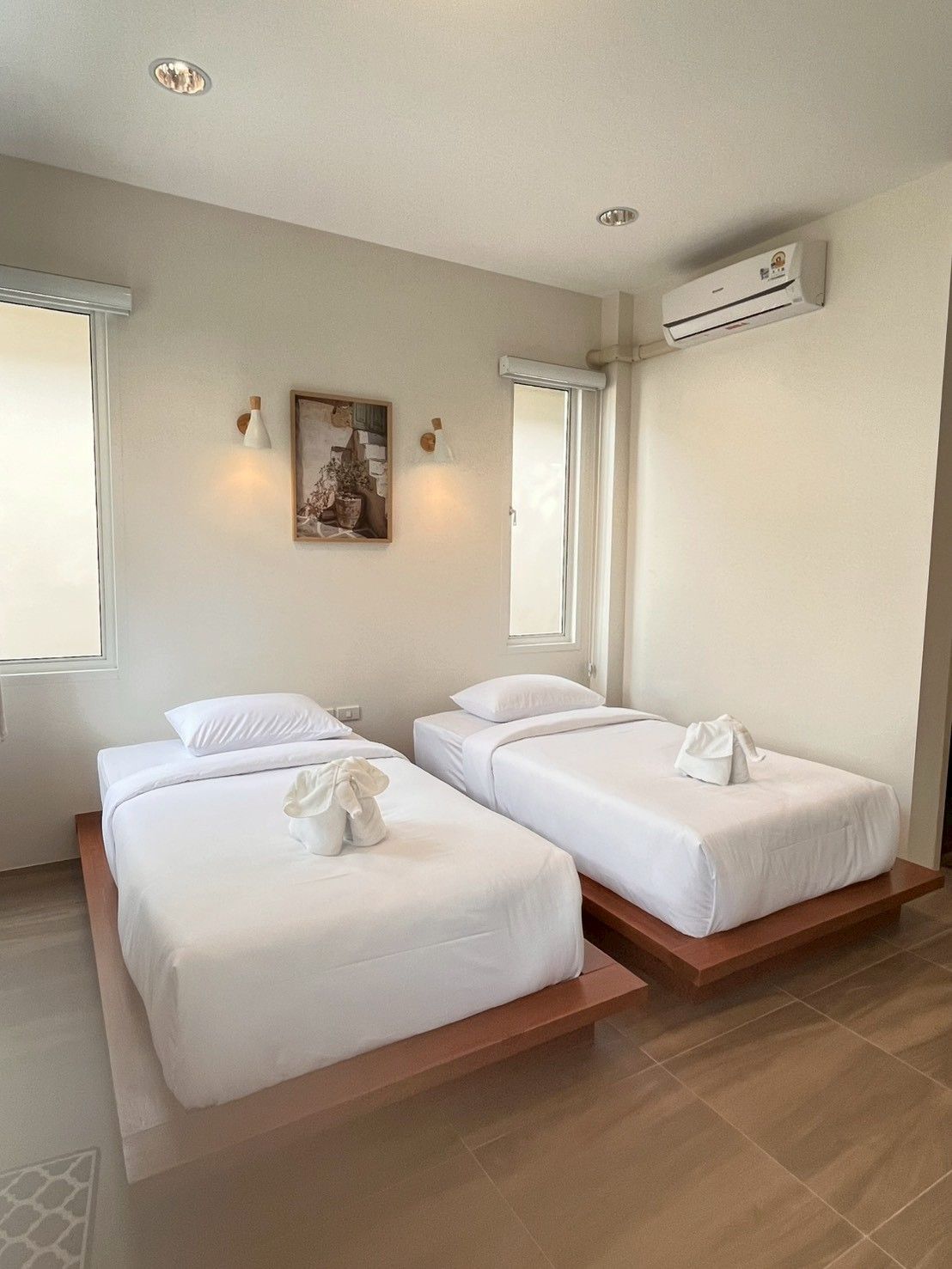 A hotel room with two beds, a painting on the wall, an air conditioning unit, and a window.