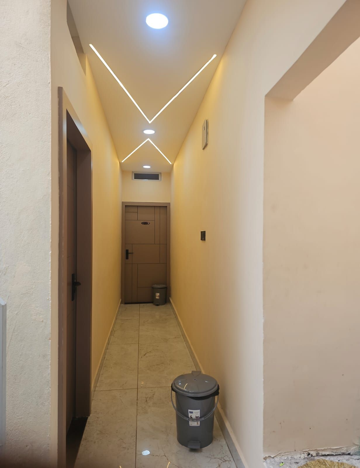 Um corredor estreito com uma porta no fim, uma lixeira no centro e um aparelho de iluminação no teto.