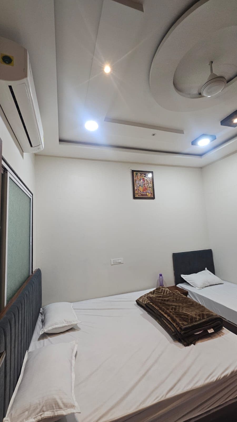 Een hotelkamer met een bed, een nachtkastje, een wandairconditioner, een plafondventilator, inbouwverlichting en een inlijstbaar schilderij aan de muur.