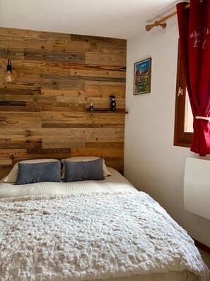 A bedroom with a wooden headboard, a bed with white bedding, two pillows, a red curtain, a wall-mounted shelf with toiletries, and a framed picture. - Le Dahut Blanc, a charming retreat at the foot of the slopes (Morillon)