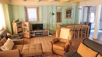 Un saló amb un sofà, una butaca i una televisió. Hi ha una taula de menjador amb caires i una finestra amb cortines.