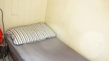 Habitació petita amb un llit individual, un matalàs gris i un coixí ratllat.