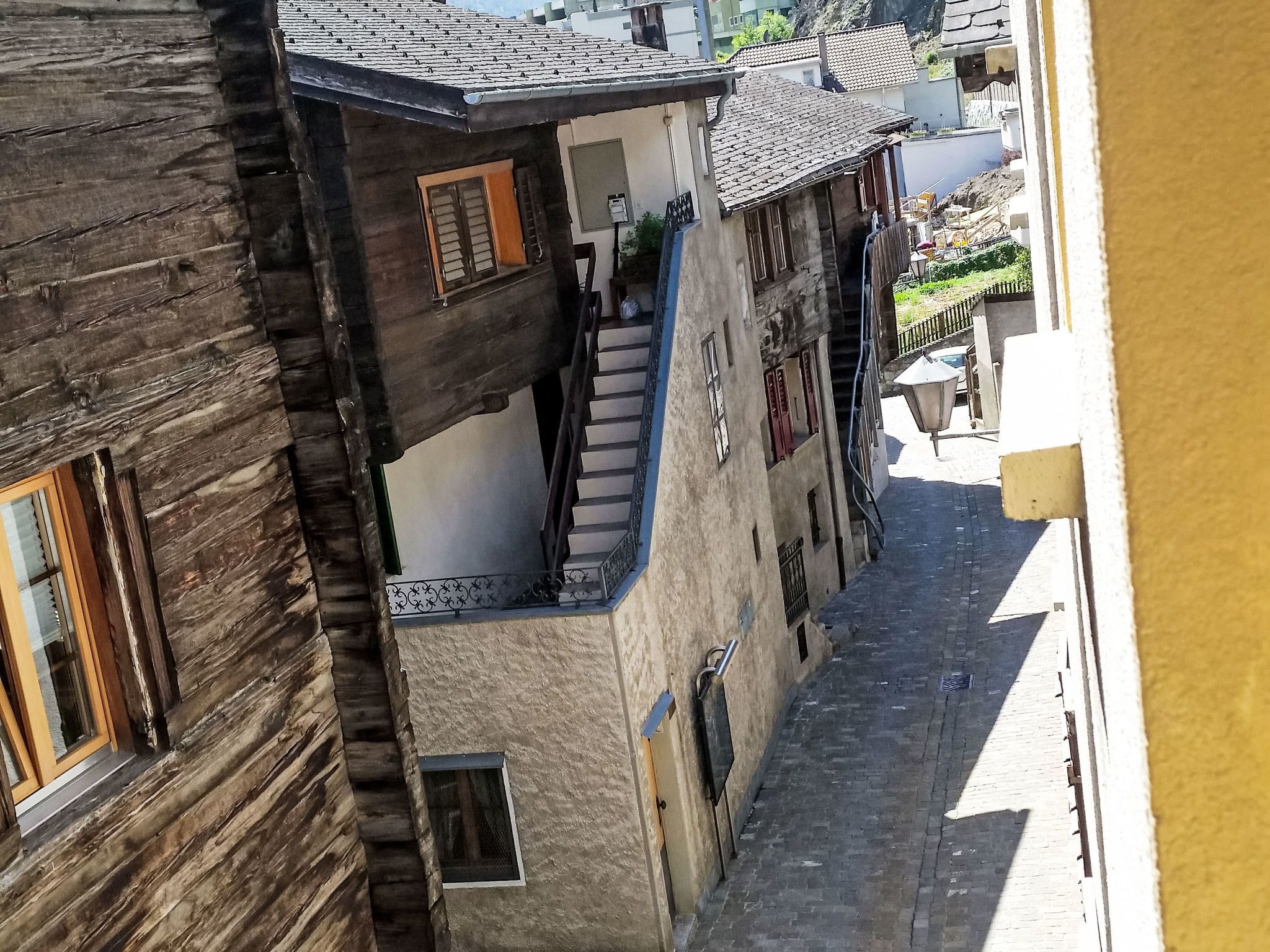 Une rue étroite, bordée de bâtiments des deux côtés, dont l’un présente une façade en bois et l’autre, un bâtiment en pierre avec un escalier apparent.