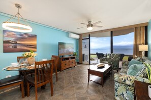 A living room with a dining area, a ceiling fan, a TV, and a view of the ocean. - Ground-Floor Oceanfront 1BD with AC & Island Views -Kahana Reef 120 (Lahaina)