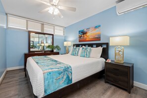 A bedroom with a bed, bedside tables, a nightstand, a ceiling fan, a painting, and a window with blinds. - Ground-Floor Oceanfront 1BD with AC & Island Views -Kahana Reef 120 (Lahaina)