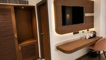 A modern hotel room with a wooden wardrobe, a mounted flat-screen TV, a built-in shelf with toiletries, and a seating area.