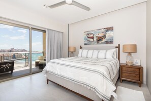 Une chambre avec un grand lit, deux lampes de chevet, une table de nuit, un tableau et une vue sur la mer, visible à travers une porte coulissante.