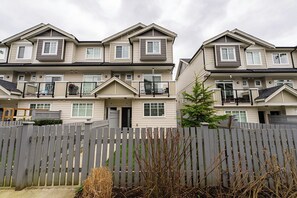 A row of two-story townhouses with balconies, a wooden fence, and a small tree in the foreground.