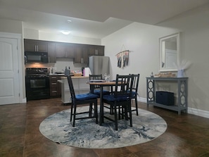 A dining area with a round table and four chairs, a kitchen with dark cabinets and stainless steel appliances, and a small side table with decorative items. - The Bluebird Inn (Fort McMurray)