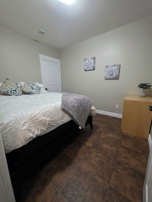 A bedroom with a bed, a door, a wall with two paintings, and a small wooden cabinet.