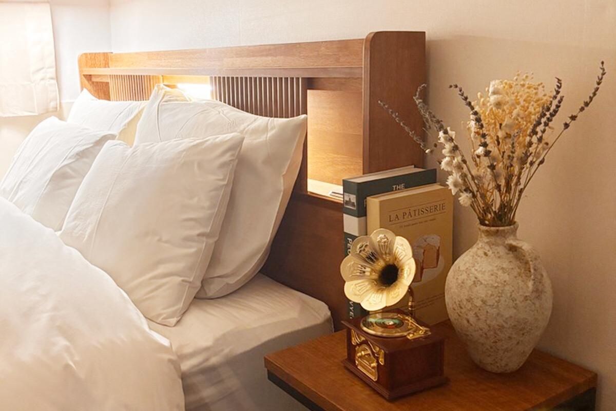 Een net opgemaakt bed met witte lakens, een nachtkastje met een boek getiteld 'The pâtisserie' en een vaas met gedroogde bloemen.