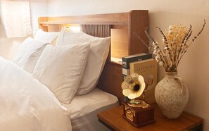 Een net opgemaakt bed met witte lakens, een nachtkastje met een boek getiteld 'The pâtisserie' en een vaas met gedroogde bloemen.