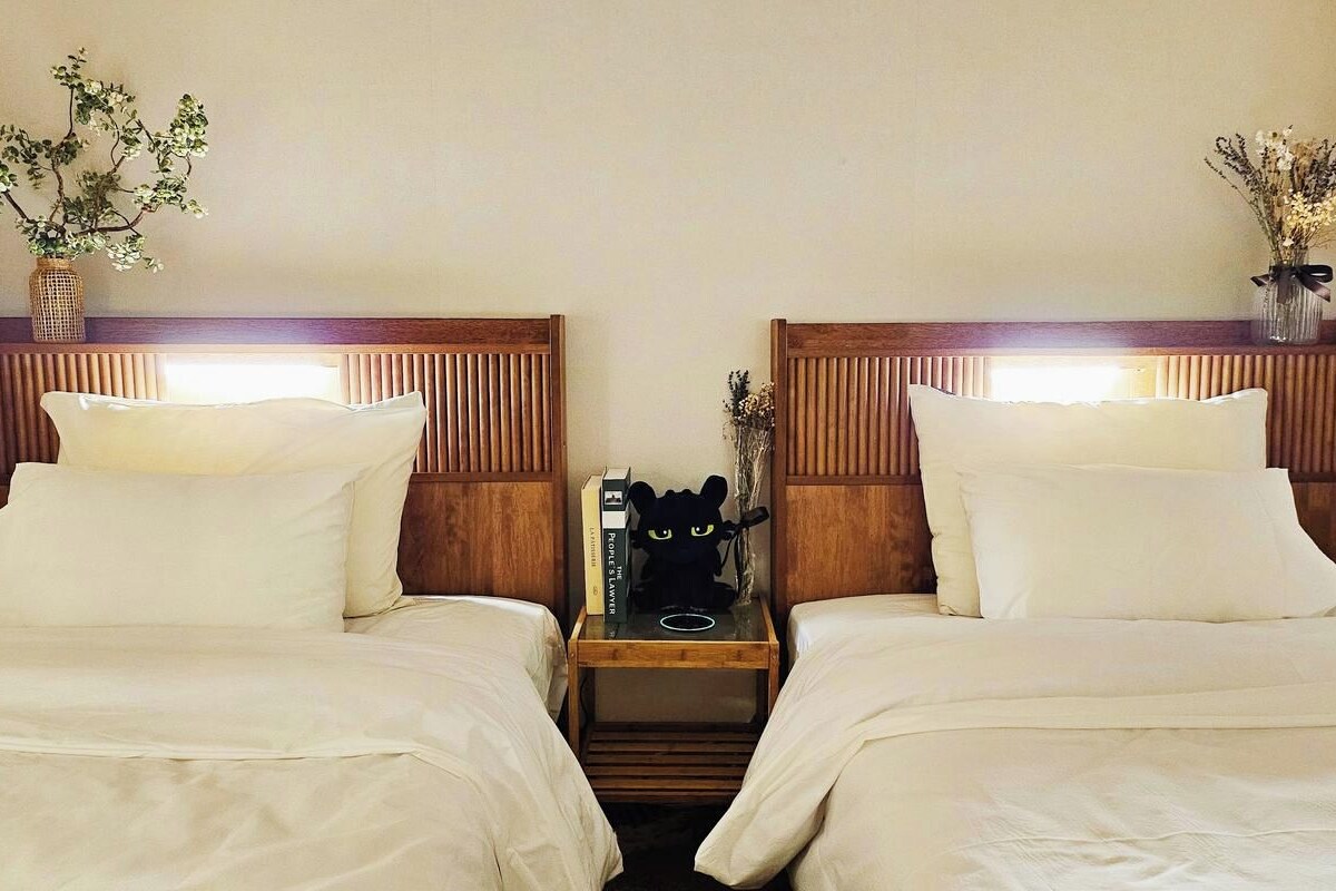 Een hotelkamer met twee bedden, houten hoofdborden, wit beddengoed, een nachtkastje met een lamp en decoratieve vazen.