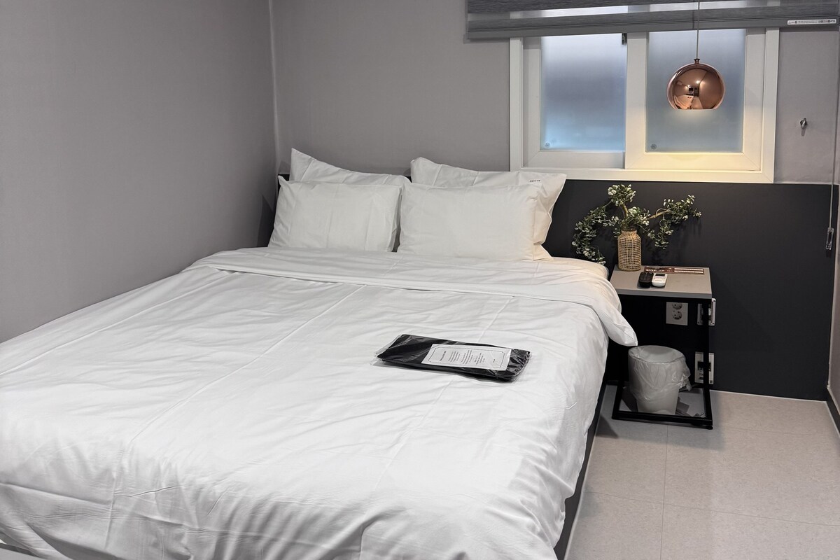 Een slaapkamer met een bed, een airconditioning, een raam met zonwering, een nachtkastje met een vaas en een hanglamp.