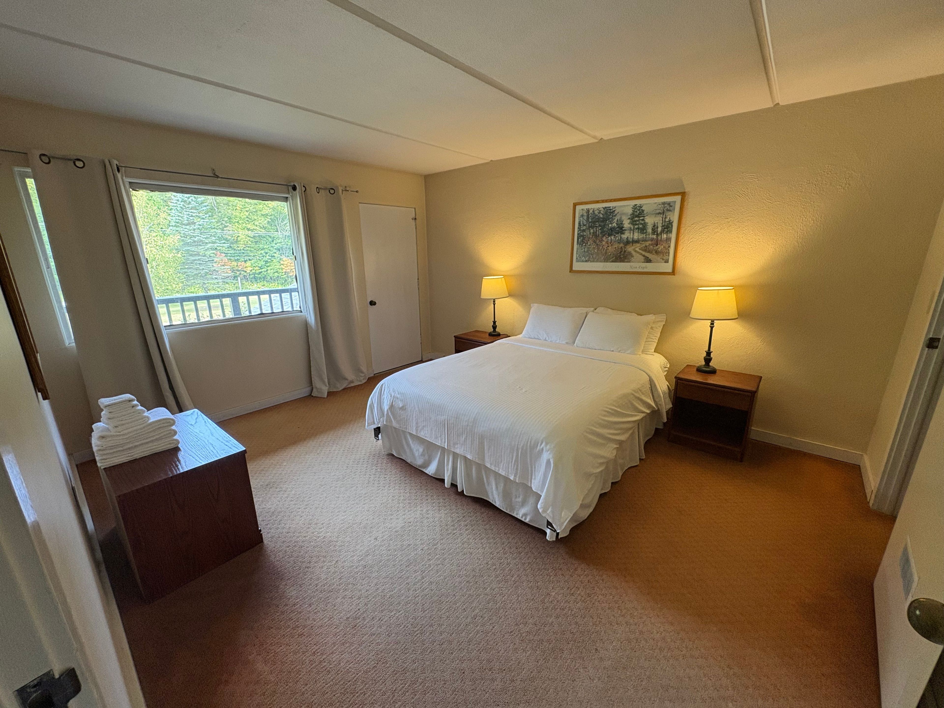 Quarto de hotel com cama, duas mesas de cabeceira com luminárias, uma porta, uma janela com cortinas e um quadro na parede.