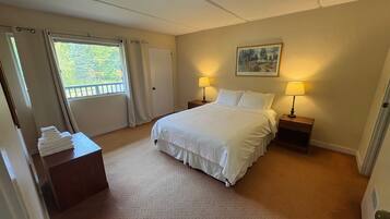 Quarto de hotel com cama, duas mesas de cabeceira com luminárias, uma porta, uma janela com cortinas e um quadro na parede.