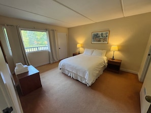 Quarto de hotel com cama, duas mesas de cabeceira com luminárias, uma porta, uma janela com cortinas e um quadro na parede.