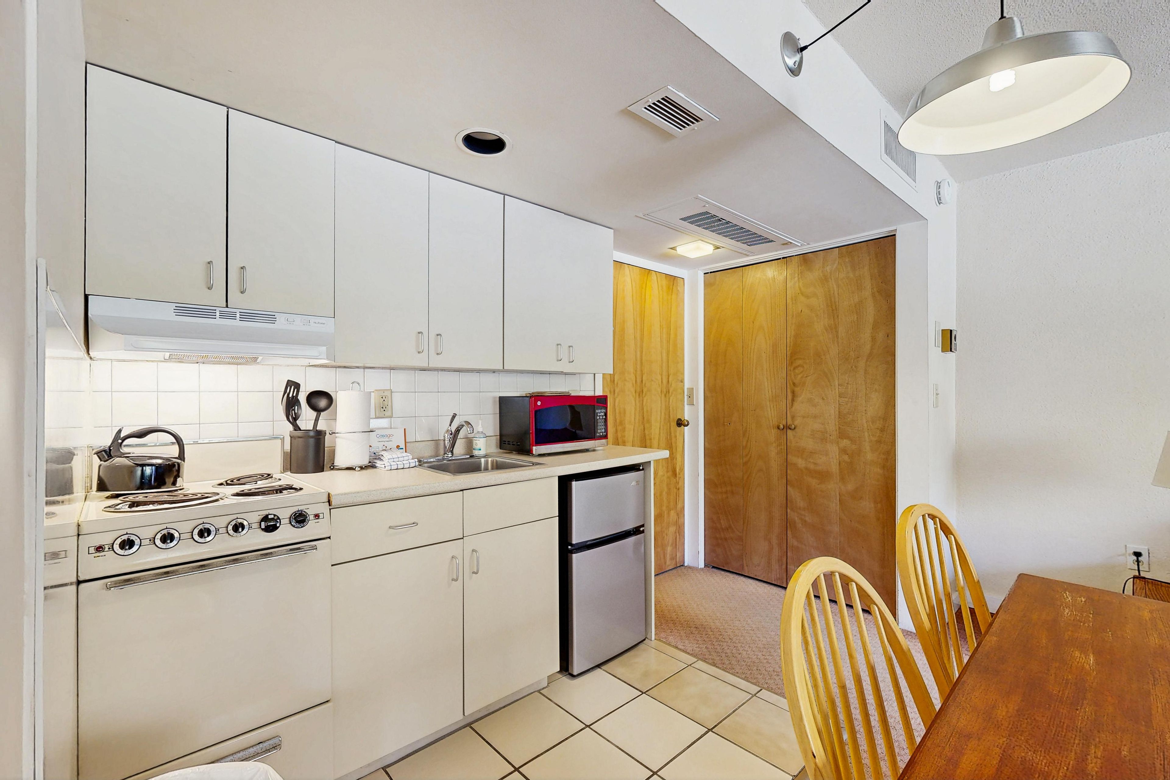 A compact kitchen with white cabinets, a stove, a microwave, and a refrigerator. There is a dining table with chairs and a wooden door in the background.