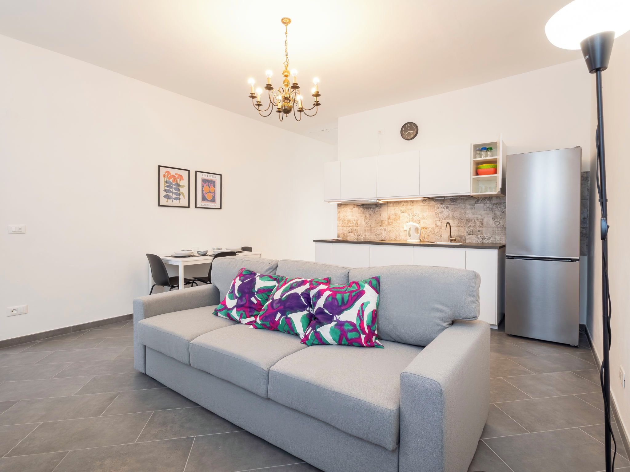 A modern living room with a grey sofa, colorful cushions, a chandelier, a dining area with a table and chairs, and a kitchen with a refrigerator and tiled backsplash.