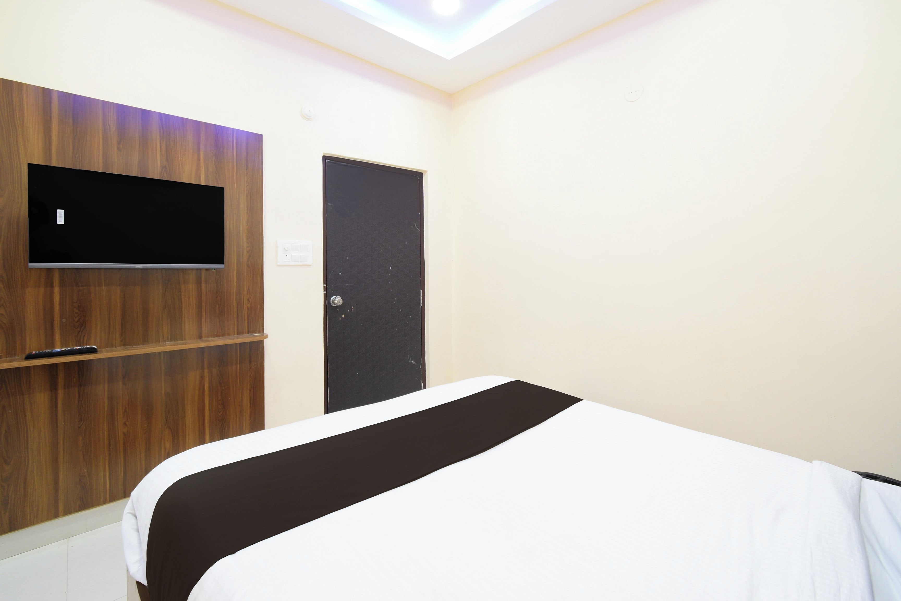 Een slaapkamer met een bed, een aan een houten paneel bevestigde televisie, een deur en een lichtschakelaar aan de muur.