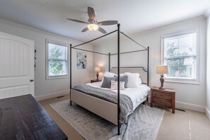 A bedroom with a four-poster bed, a ceiling fan, two bedside tables with lamps, a bench, and a large window with blinds. - 3BR home in Reynolds Landing with patio, grill & foosball (Greensboro)