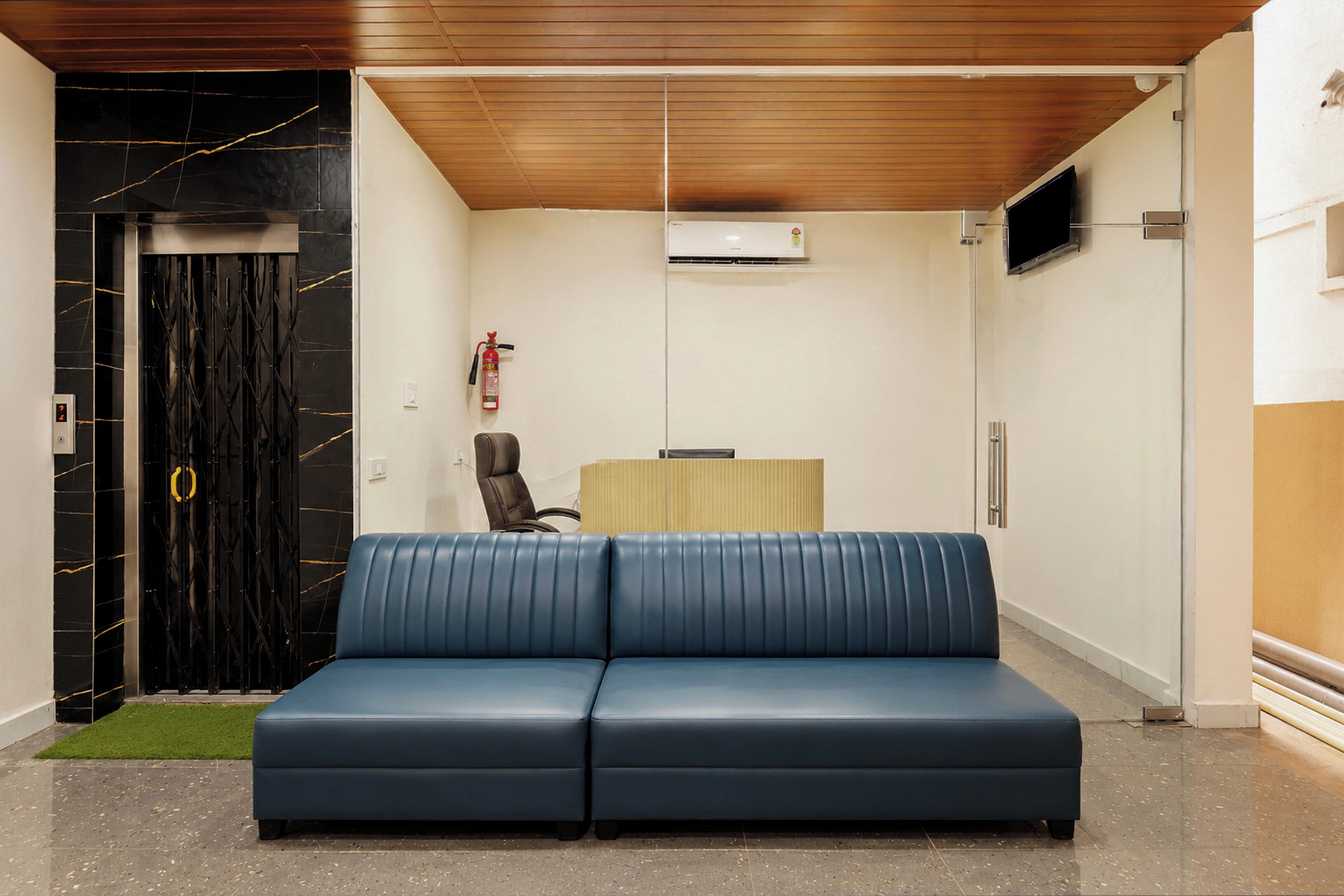 A modern waiting area with a blue sofa, a fire extinguisher, an air conditioning unit, and a closed door with a keypad.