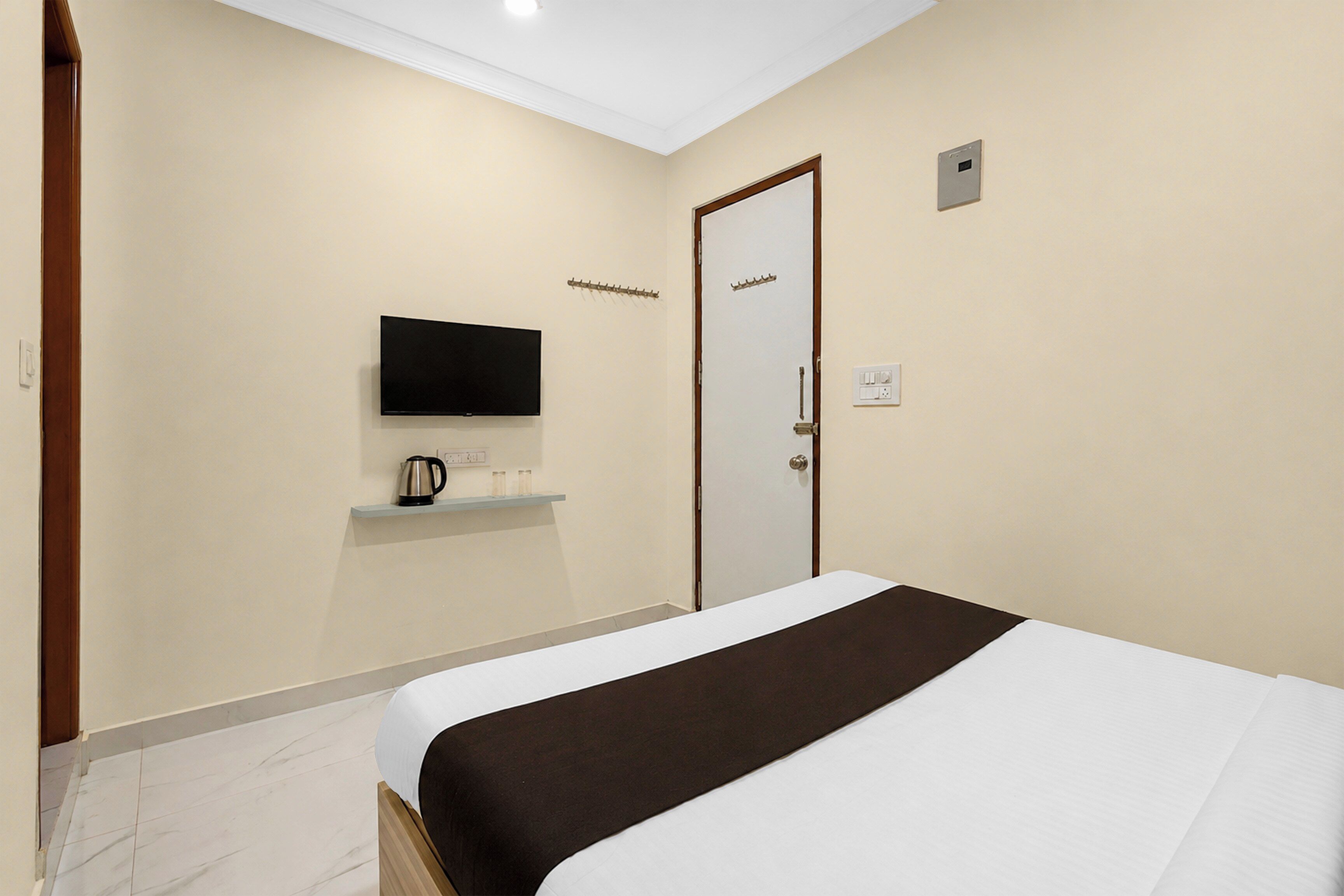 A hotel room with a bed, a television mounted on the wall, a door with a coat hook, and a small shelf with a kettle.