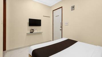 A hotel room with a bed, a television mounted on the wall, a door with a coat hook, and a small shelf with a kettle.
