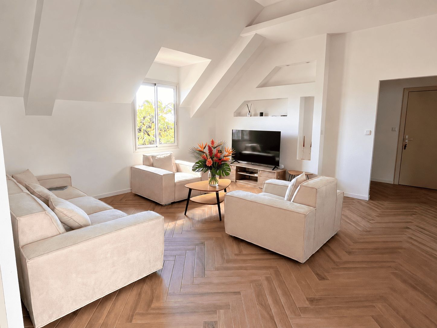 Un salon moderne avec un parquet en chevrons, un téléviseur à écran plat et une grande fenêtre donnant sur la forêt.