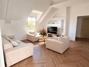 A modern living room with a herringbone wood floor, a flat-screen TV, and a large window with a view of trees.