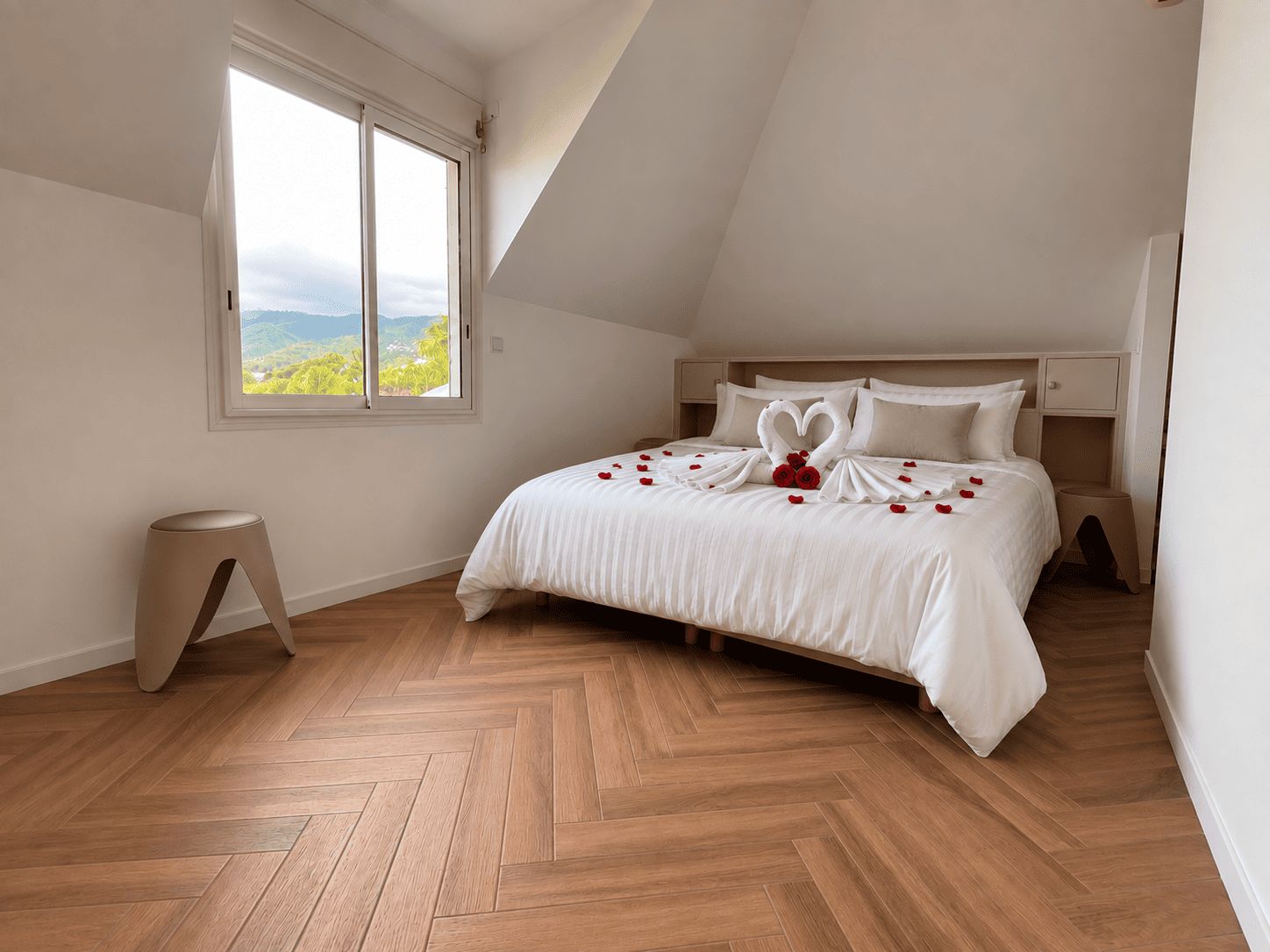 Une chambre avec une grande fenêtre, un lit recouvert de draps blancs et un parquet en bois disposé en chevrons.