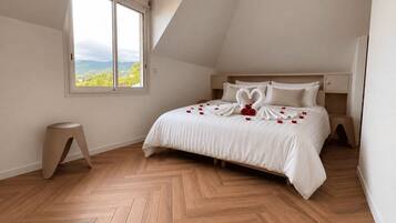 Un dormitorio con un ventanal grande, una cama con ropa de cama blanca y un suelo de madera en espiga.