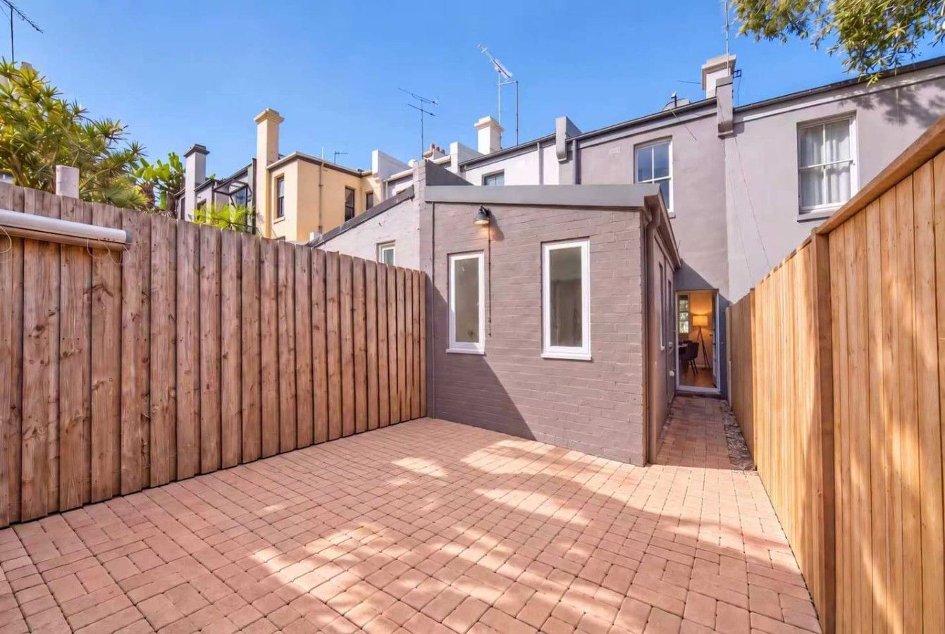Une cour entourée d’une clôture en bois, avec une terrasse en briques et un petit bâtiment doté de fenêtres.