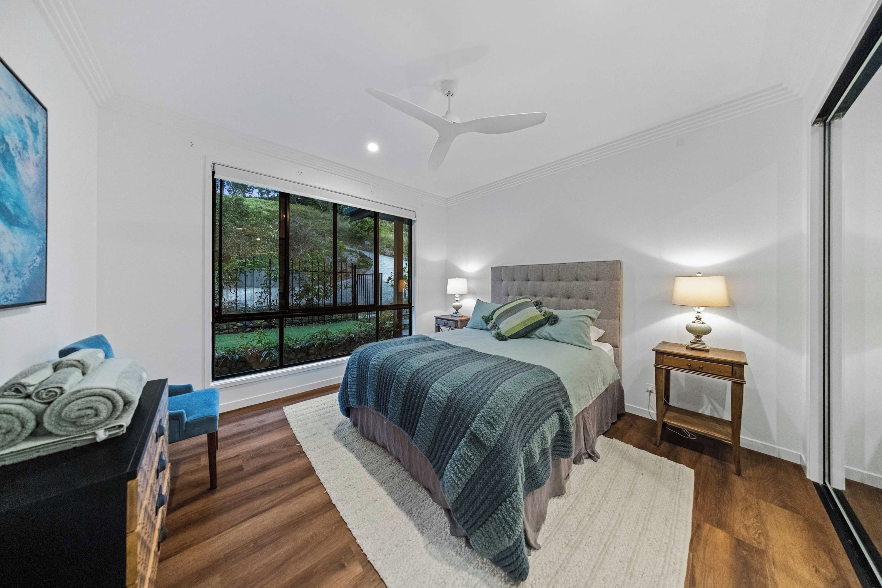 Ein Schlafzimmer mit großem Fenster, einem Bett mit grauem Kopfteil, zwei Nachttischen mit Lampen, Holzboden und Deckenventilator.