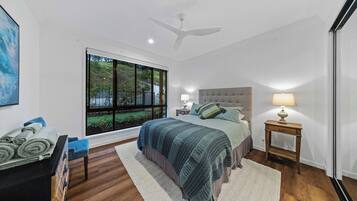 Ein Schlafzimmer mit großem Fenster, einem Bett mit grauem Kopfteil, zwei Nachttischen mit Lampen, Holzboden und Deckenventilator.
