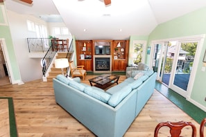 A spacious living room with a large blue sofa, wooden flooring, a staircase, and a kitchen area with wooden cabinets. - Soundside- Private Pool, Comm Amenities-MS842, Bella Vita OBX (Corolla)